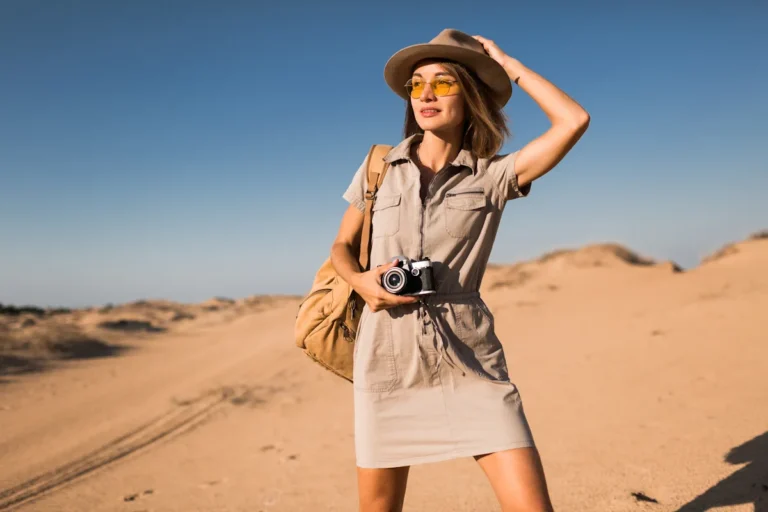 Une femme en robe beige avec un chapeau, des lunettes de soleil et un sac ainsi qu'un appareil photo au milieu du desert