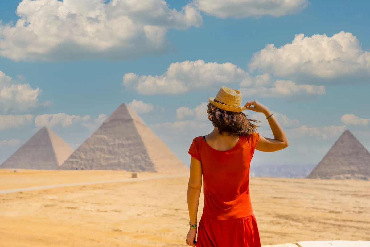 Une femme en robe et avec un chapeau qui regarde de loin les pyramides