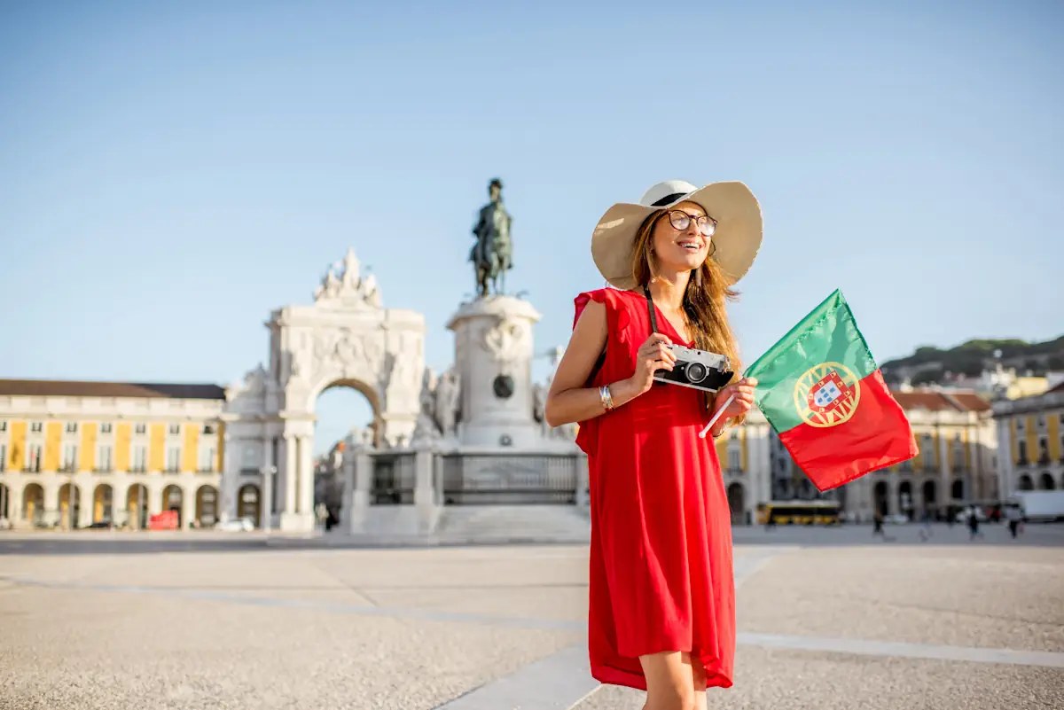 Une femme en robe rouge et avec un chapeau de paille avec un appareil photo et un drapeau sur une place de Lisbonne