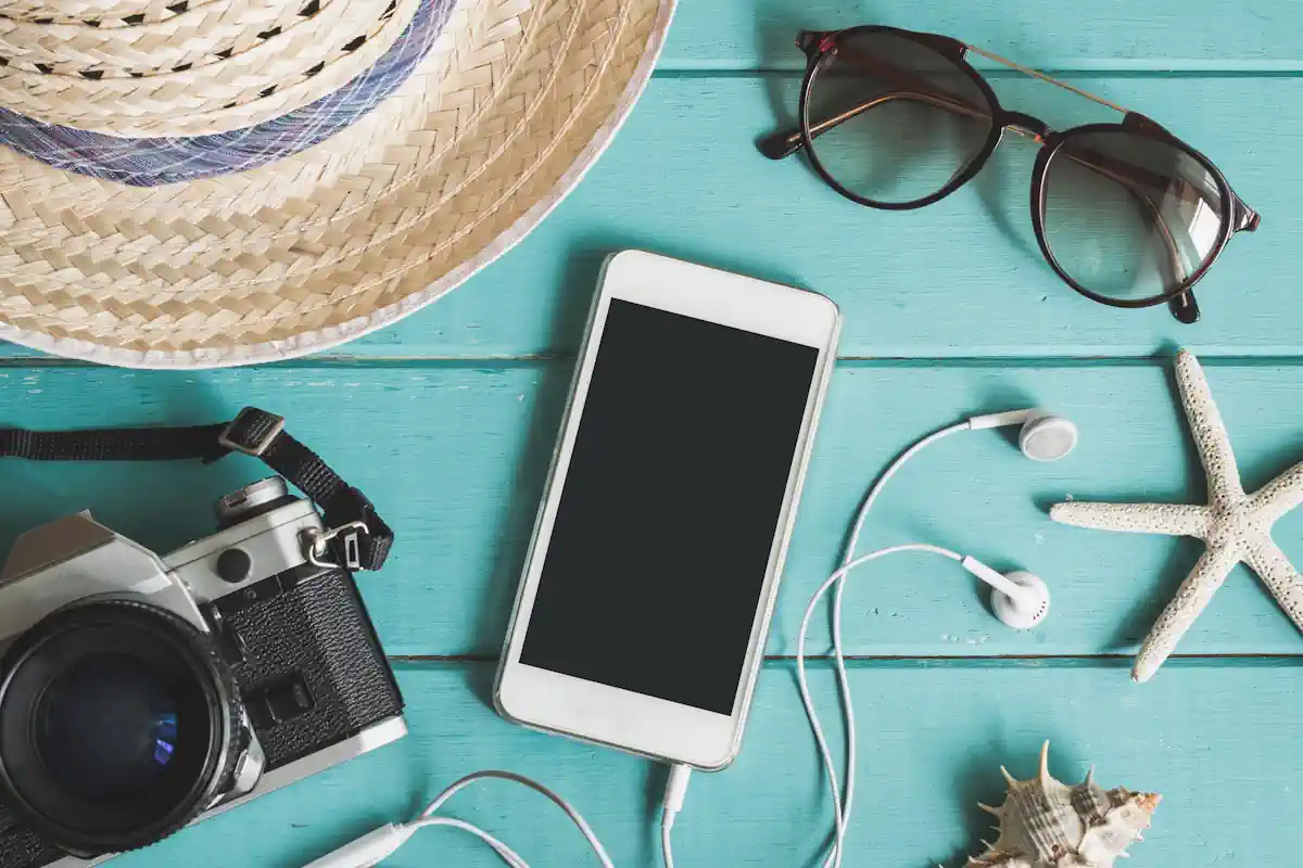 Un smartphone avec des écouteurs filaires, posé à côté d'un appareil photo, d'une paire de lunettes de soleil et d'un chapeau sur une surface bleue